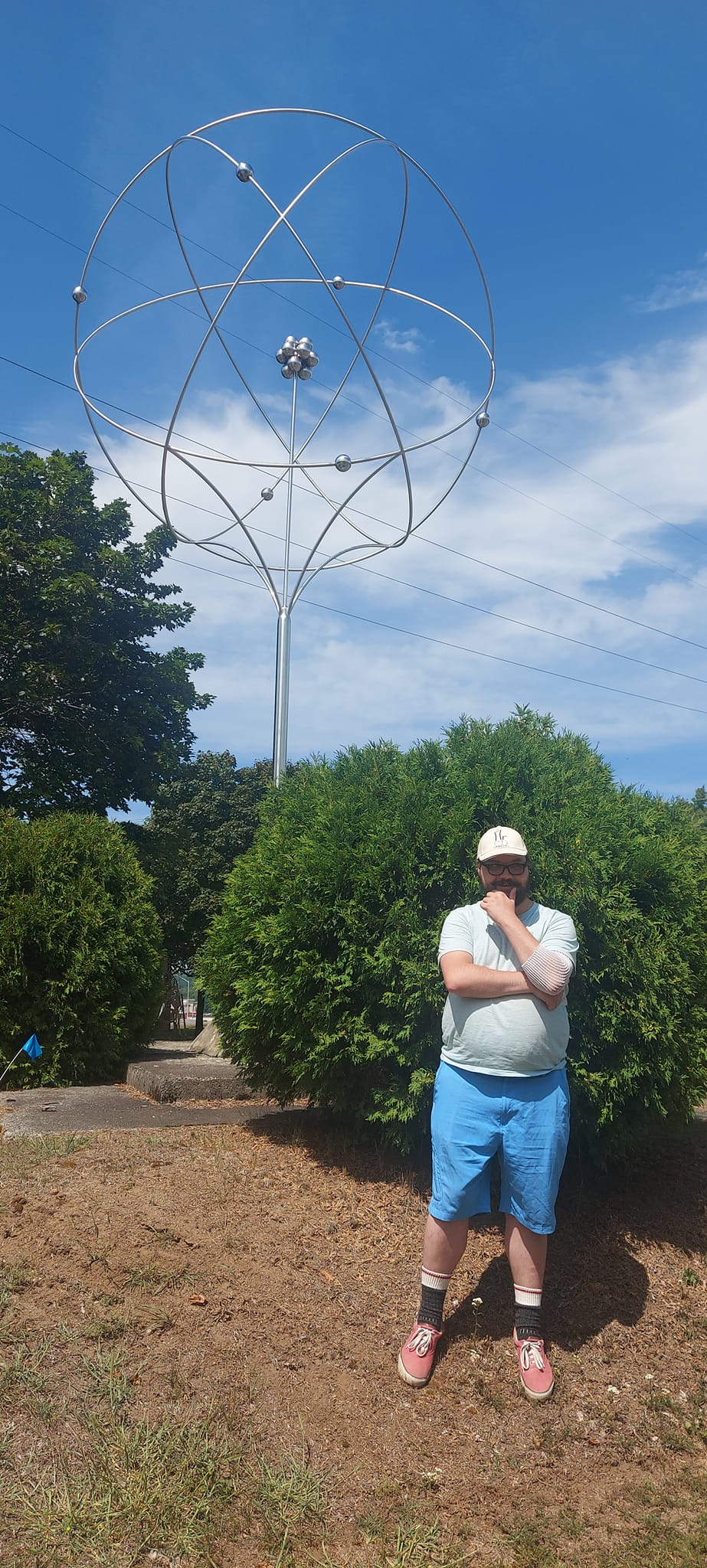 A man short hair and bushy facial hair stands in front of a giant atom sculpture, scratching his chin ponderously